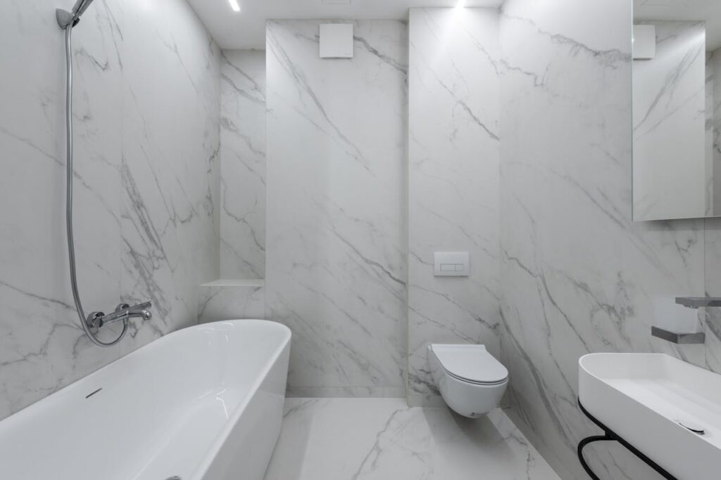 Interior of stylish bathroom with marble walls and bath placed near toilet and ceramic sink in light room with mirror